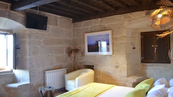 Un dormitorio con paredes de piedra, una cama, un sillón blanco, una mesita y una chimenea.