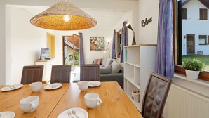 Ein Essbereich mit einem Holztisch, gedeckt für vier Personen, einer Korbtreleuchte und Blick auf ein Haus durch ein Fenster mit violetten Vorhängen.