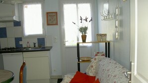 A kitchen with a stove, sink, and a dining table with chairs. There is a sofa with floral upholstery and two orange cushions. A potted plant is on a shelf above the sofa. - Attached ground floor apartment on Enclosed grounds (Plouay)