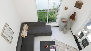 A modern living room with a grey sofa, a coffee table with magazines, and a view of a garden through a large window. - Design House in a famous area - St. Sebastian Curch & Temple road (Negombo/Katuwapitiye)