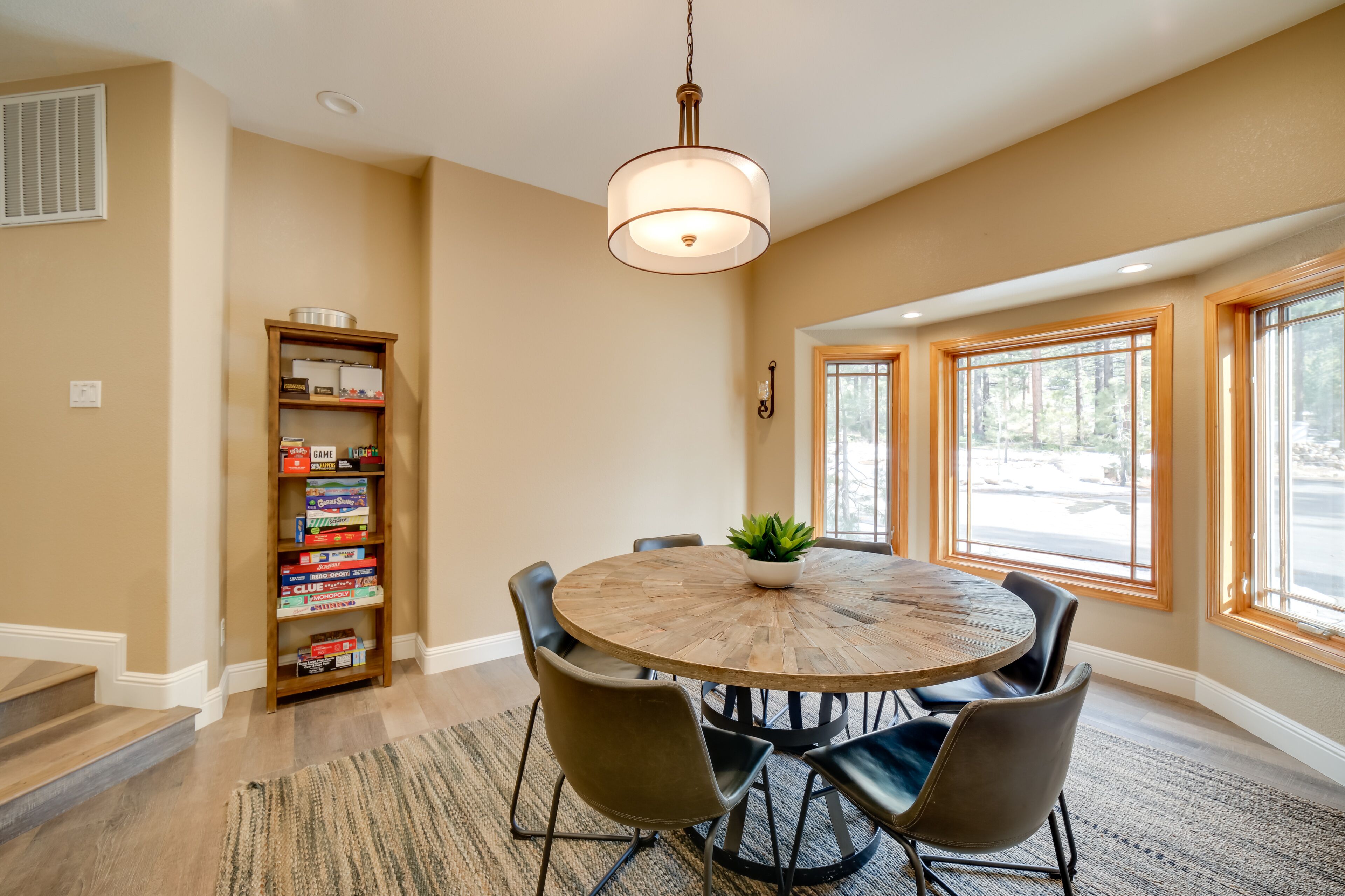 A dining area with a round wooden table, six chairs, a hanging light fixture, a bookshelf with board games, and large windows with a view of trees.