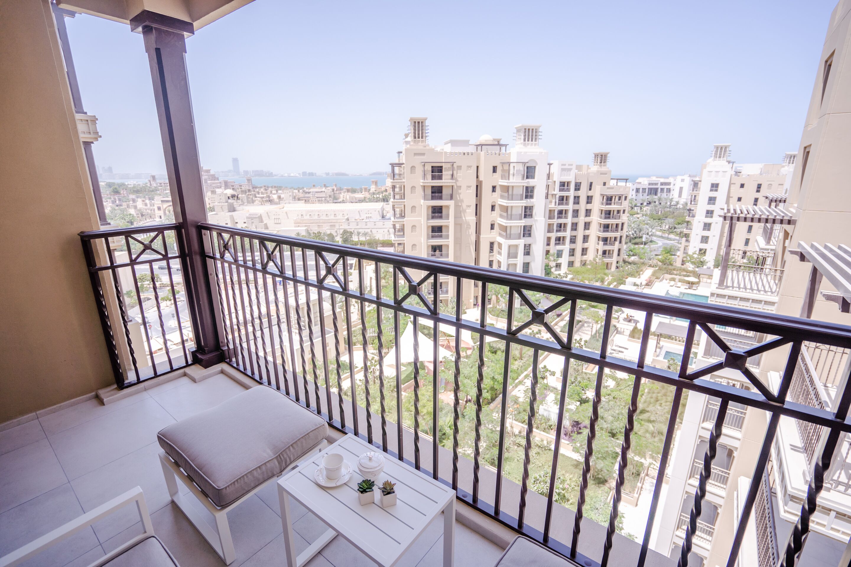A balcony with a white cushioned bench, a small table with a cup and two small potted plants, and a view of surrounding buildings.