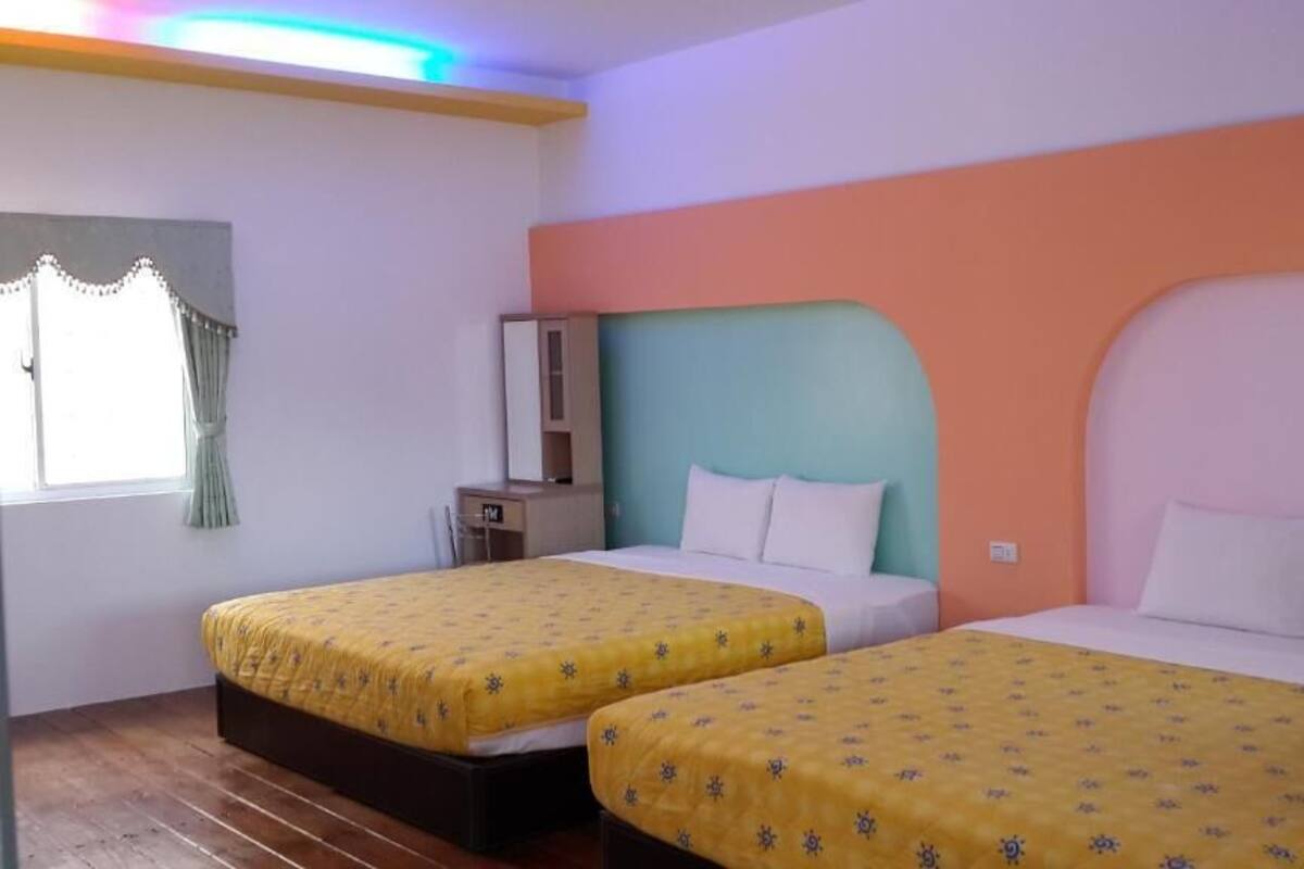 Quarto de hotel com duas camas, cada uma com colcha amarela, uma mesa de cabeceira, um espelho e uma janela com cortinas.