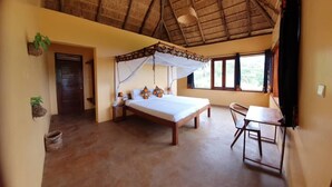 A room with a bed under a thatched roof, a small table, and a chair. - Kikonko Lodge (Buseruka)
