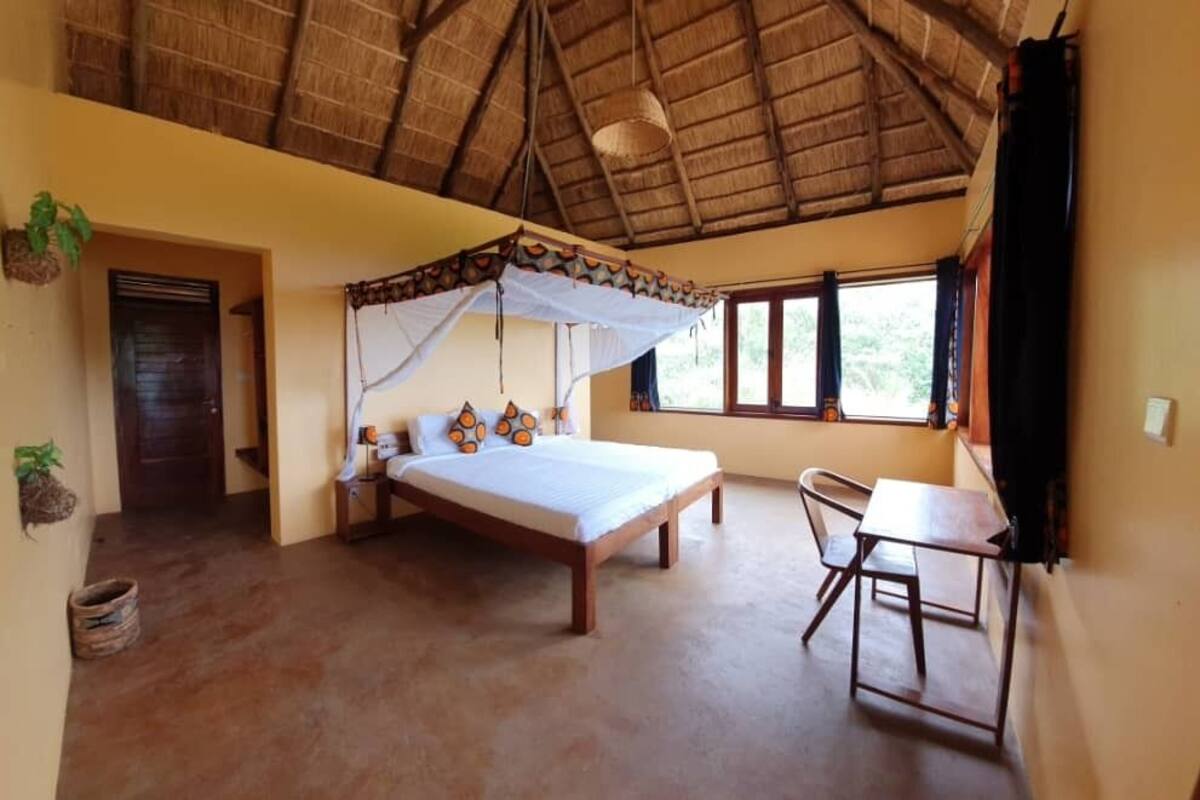 A room with a bed under a thatched roof, a small table, and a chair.