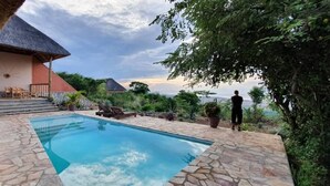 A swimming pool with stone tiles, a thatched-roof building, and a view of the ocean. - Kikonko Lodge (Buseruka)