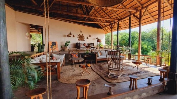 An open-plan living area with a wooden ceiling, a sofa set, a dining table, and a view of lush greenery. - Kikonko Lodge (Buseruka)