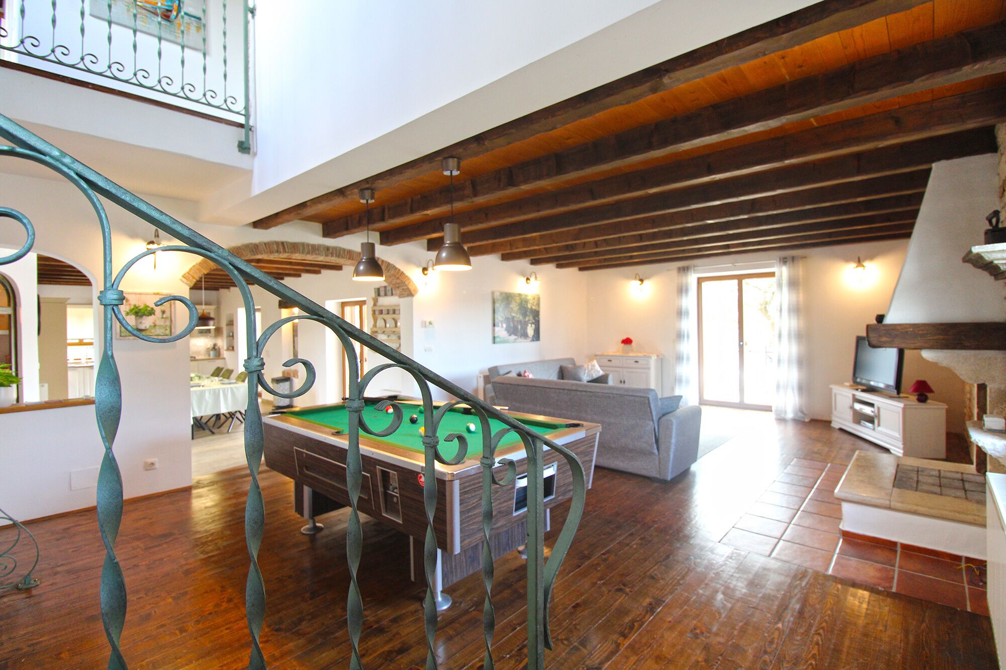 Un vaste espace de vie comprenant une table de billard, un canapé et un escalier avec des rampes en fer forgé.