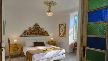 Un dormitorio con una cama grande, dos mesitas de noche con lámparas, un cabecero decorativo, un radiador y una ventana con vidrios verdes.