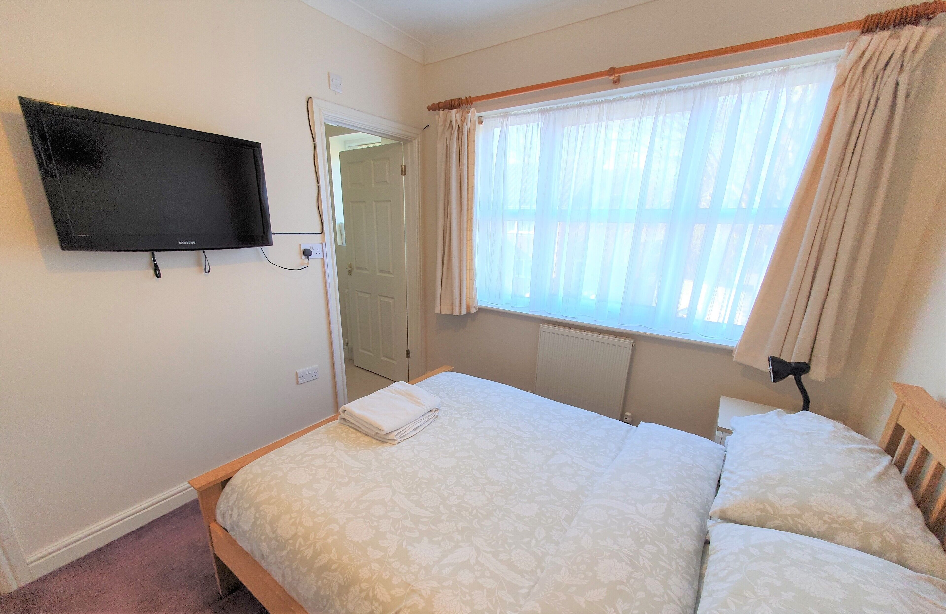 A bedroom with a bed, a television mounted on the wall, a window with curtains, and a radiator.