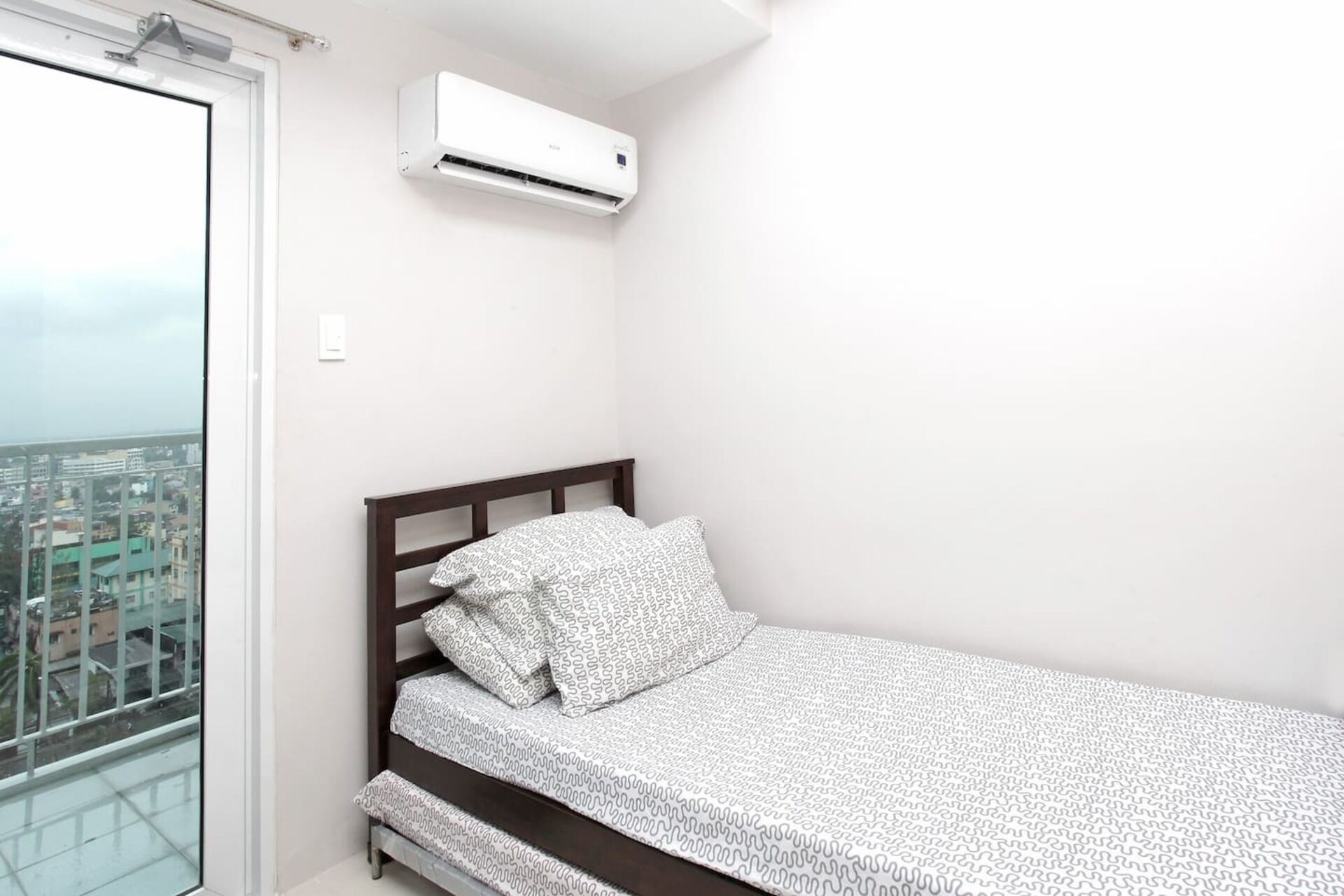 A small, single-bed room with a city view, white walls, and a mounted air conditioner.