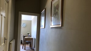 Een gang met een deur aan de linkerkant, een klok aan de muur en een tafel met stoelen op de achtergrond.