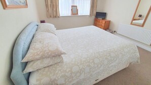A neatly made bed with a floral-patterned bedspread and pillows, a dresser with a television, a window with curtains, and a radiator.