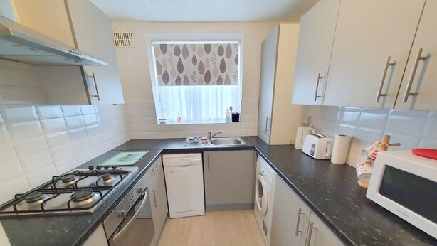 A compact kitchen with a gas stove, dishwasher, microwave, and a small window with a leaf-patterned curtain.