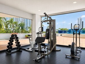 A gym with various exercise equipment, including a bench press, dumbbells, and a stationary bike, with a view of palm trees and buildings.