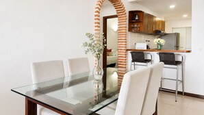 Un comedor con una mesa y sillas de vidrio, un pilar de ladrillo, una cocina al fondo y un jarrón con flores.