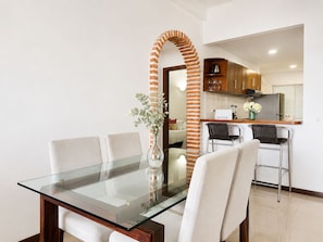 Un comedor con una mesa y sillas de vidrio, un pilar de ladrillo, una cocina al fondo y un jarrón con flores.