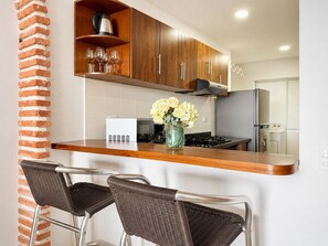 A kitchen with a brick column, wooden cabinets, a stainless steel refrigerator, and a bar area with wicker chairs.