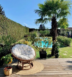 Una terraza de madera con una silla blanca acolchada, una planta pequeña en maceta y una piscina al fondo.