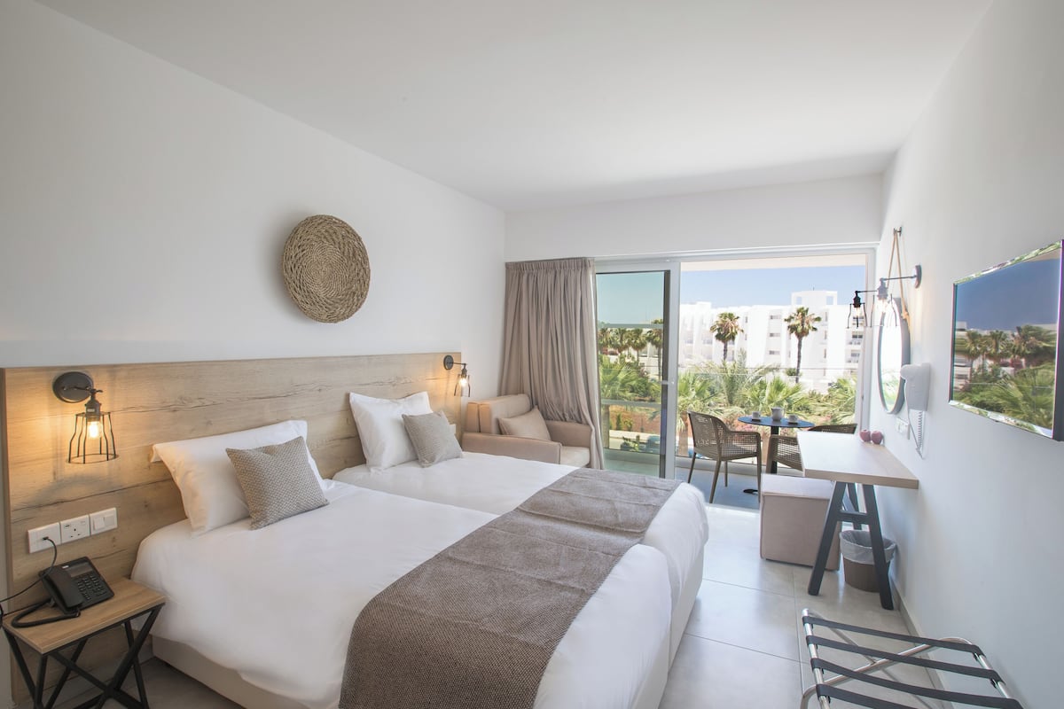 A hotel room with two beds, a desk, and a balcony with a view of palm trees and buildings.
