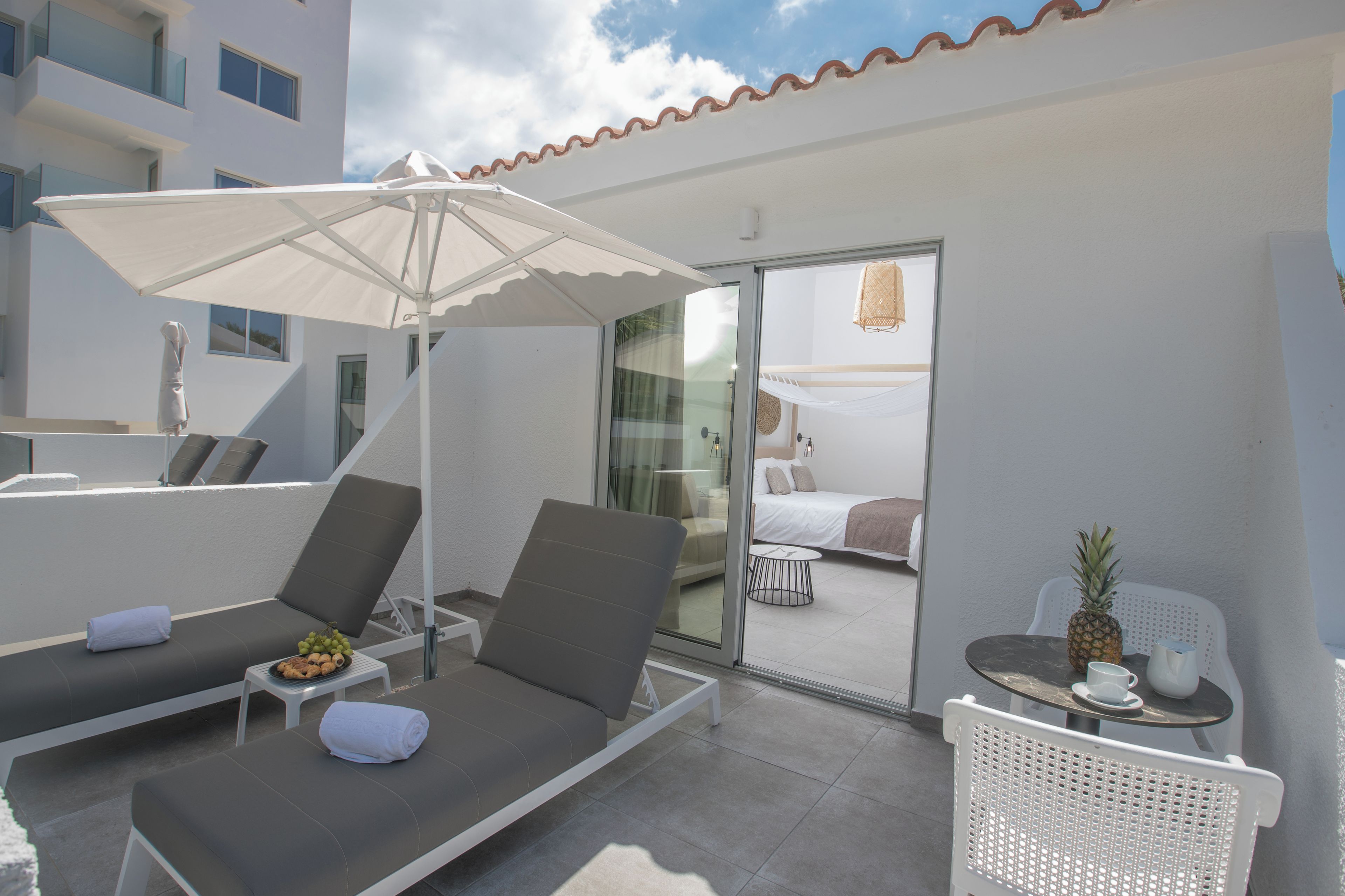 A modern hotel balcony with two lounge chairs, a table with a pineapple, and a glass door leading to a bedroom.