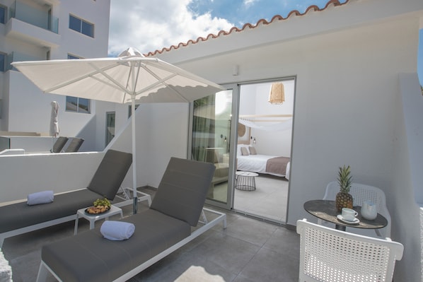 A modern hotel balcony with two lounge chairs, a table with a pineapple, and a glass door leading to a bedroom. - Bohemian Gardens Hotel (Paralimni)