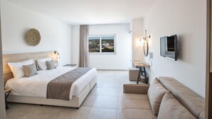 A modern hotel room with a large bed, a sofa, a TV, and a window with a city view. - Bohemian Gardens Hotel (Paralimni)