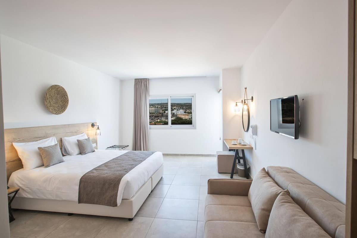 A modern hotel room with a large bed, a sofa, a TV, and a window with a city view.