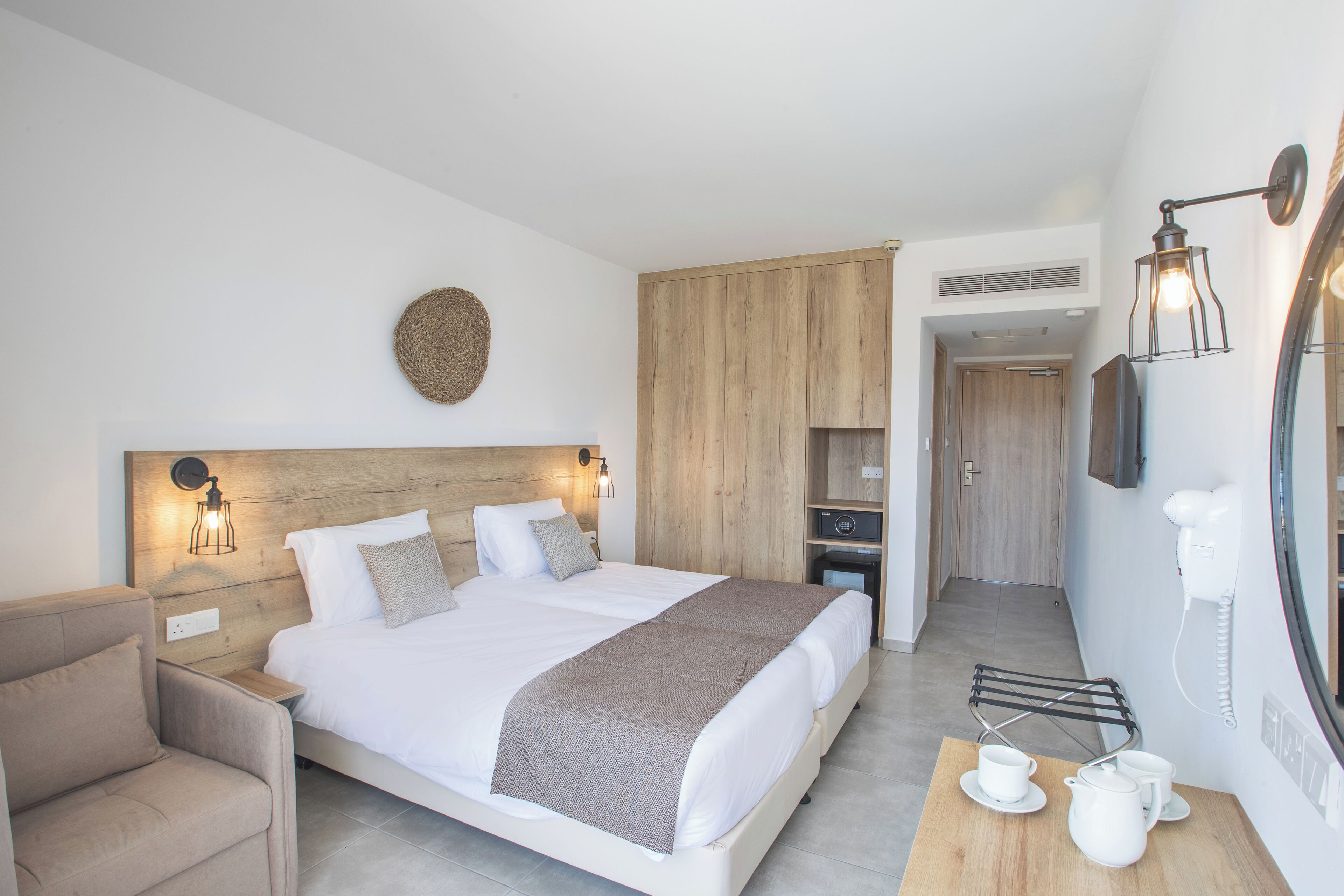 A modern bedroom with a large bed, bedside tables, a chair, a wooden wardrobe, a mirror, and a small table with cups and a pitcher.