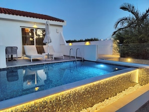 A swimming pool with lounge chairs and a covered seating area, surrounded by a white building with a tiled roof. - Bohemian Gardens Hotel (Paralimni)