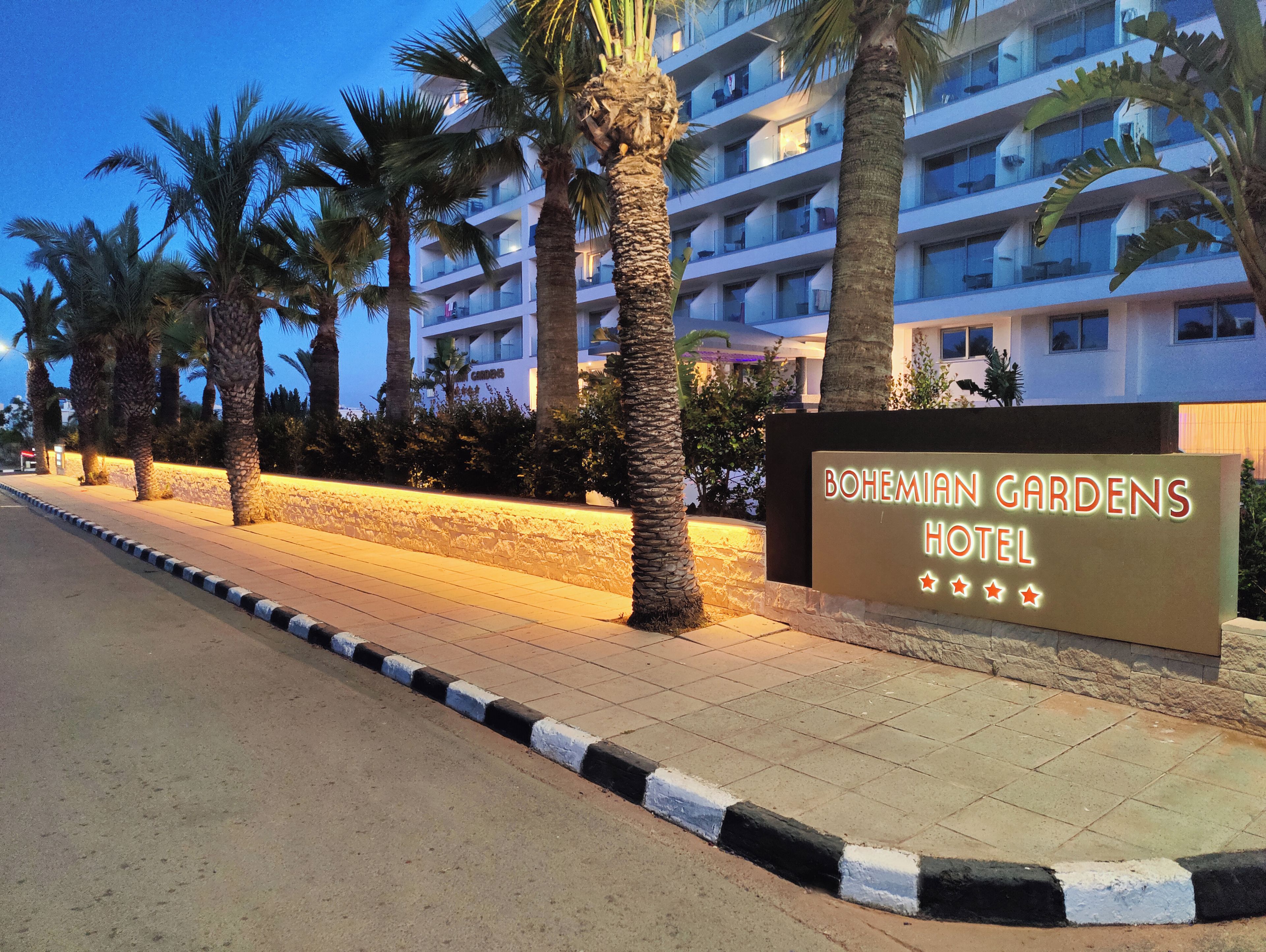 L’entrée d’un hôtel, avec une enseigne indiquant « Bohemian Gardens Hotel », des palmiers et un bâtiment doté de balcons.
