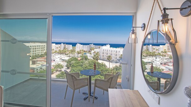 A balcony with a view of the city and ocean, a round table with chairs, and a mirror with lights. - Bohemian Gardens Hotel (Paralimni)