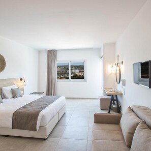 A modern hotel room with a large bed, a sofa, a small table, a TV, and a window with a city view. - Bohemian Gardens Hotel (Paralimni)