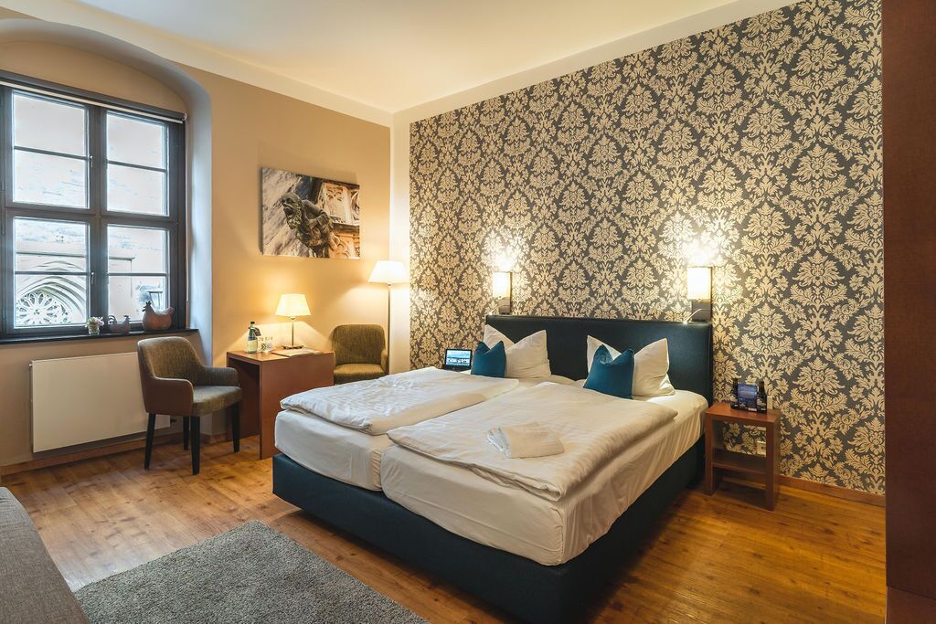 A double bed with two pillows, a nightstand with a lamp, a chair, and a window with a view of a building.