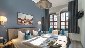 Habitación de hotel con una cama grande, dos mesitas de noche, un escritorio, una silla, un cuadro en la pared y una ventana con cortinas.