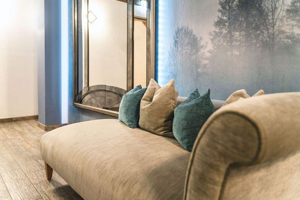 A room with a beige sofa, teal and beige cushions, a large window with a view of trees, and a mirror reflecting the room.
