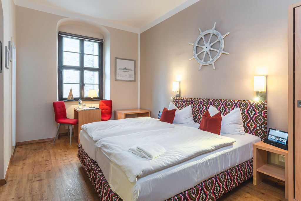 A hotel room with a large bed, a desk with a chair, a ship's wheel on the wall, and a window.