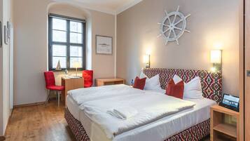 Habitació d'hotel amb un llit gran, una taula de treball amb cadira, una roda de vaixell a la paret i una finestra.