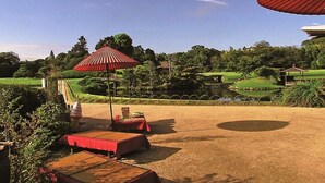 An outdoor seating area with tables and umbrellas, a landscaped garden, and a pond in the background. - Three Wellness (Okayama)