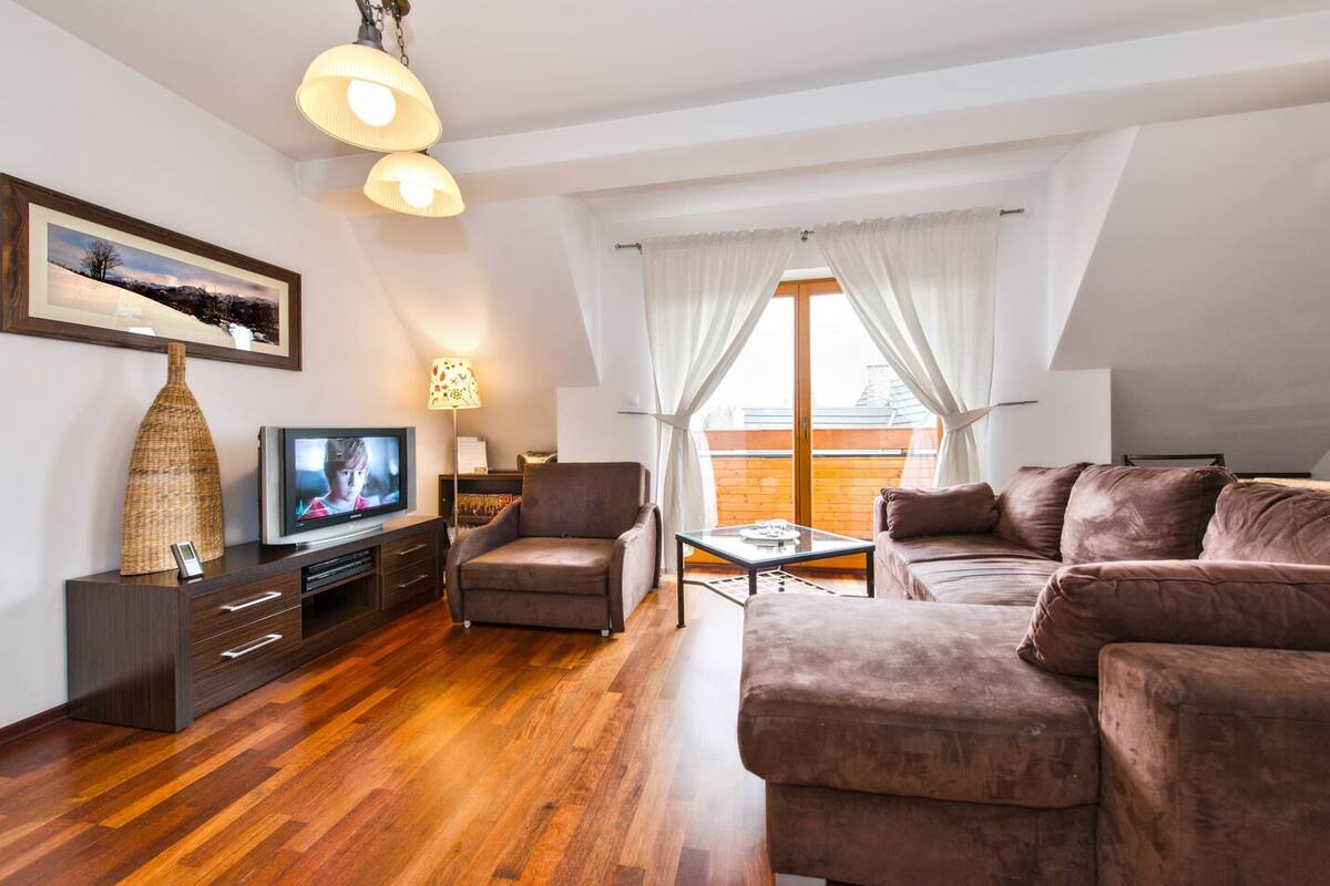 A modern living room with a wooden floor, a brown sofa, a brown armchair, a glass coffee table, a TV on a wooden cabinet, and a large window with white curtains.