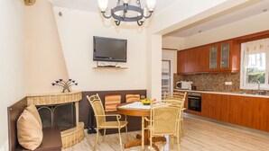 Un comedor con una mesa redonda y sillas de mimbre, un televisor montado en la pared, y armarios de madera en la cocina contigua.