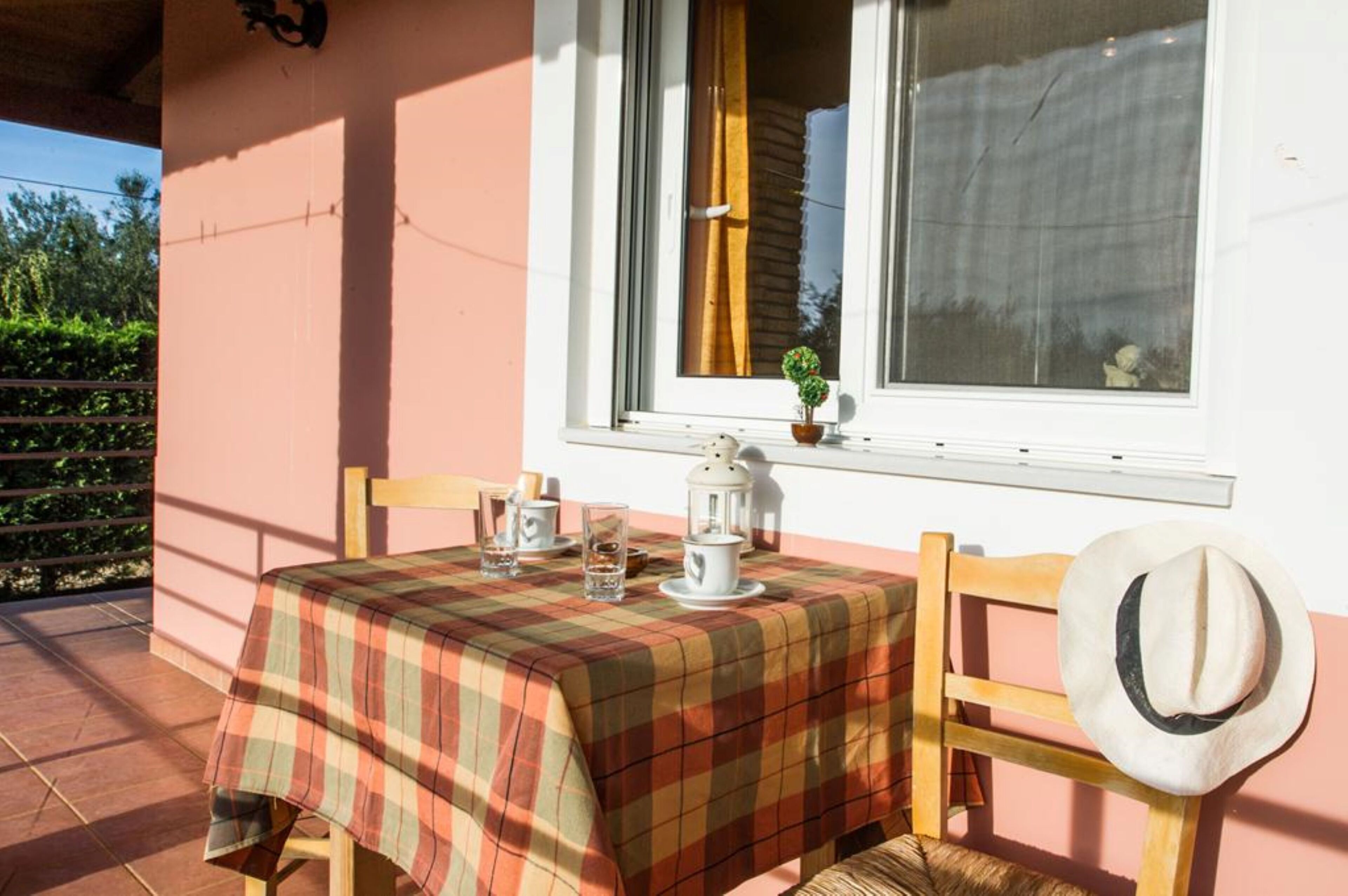 A table set with a plaid tablecloth, a white lantern, and a hat on a chair outside a building with a window.