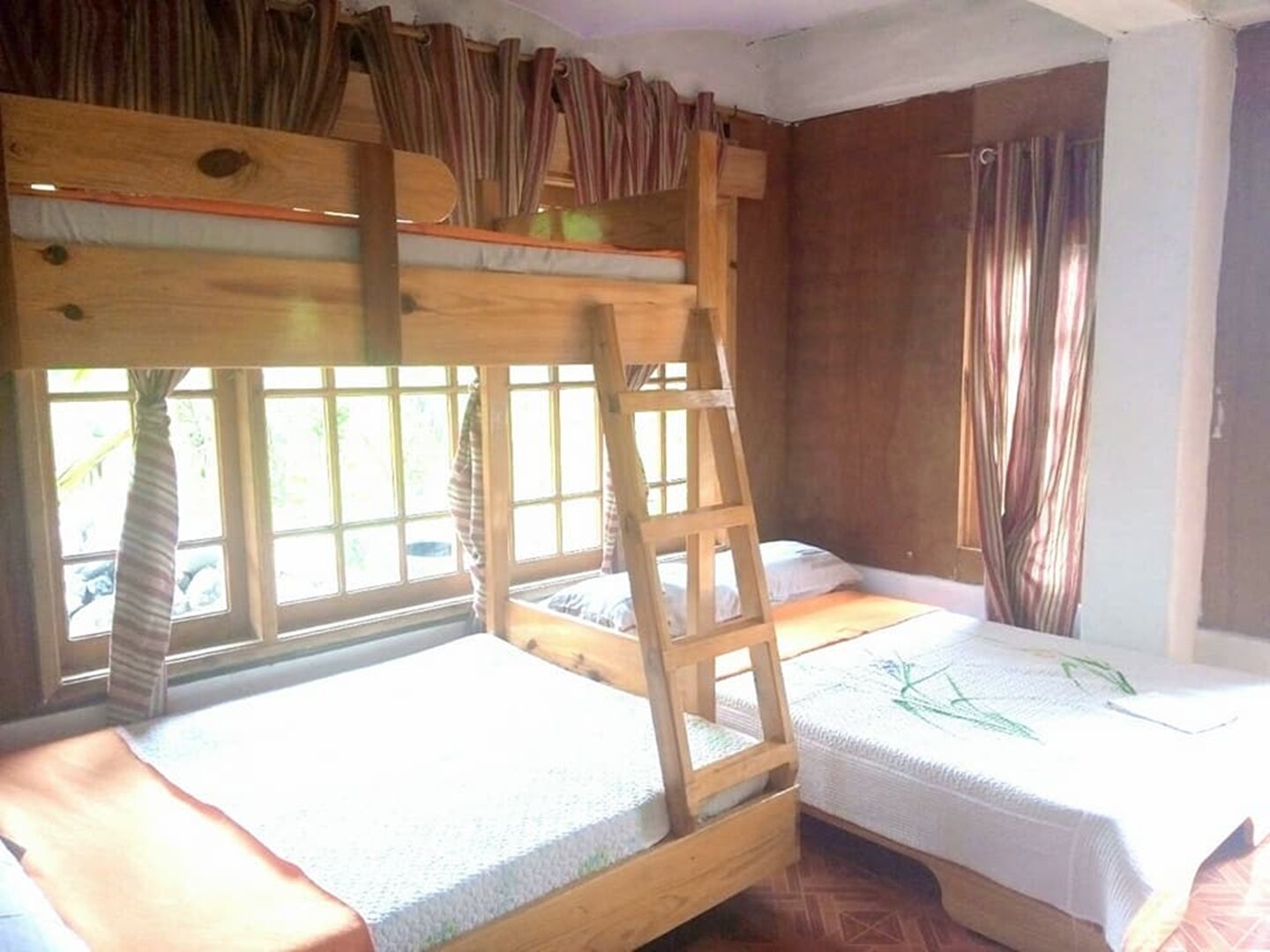 Une chambre avec un lit superposé, une échelle en bois, deux lits et une fenêtre avec des rideaux.