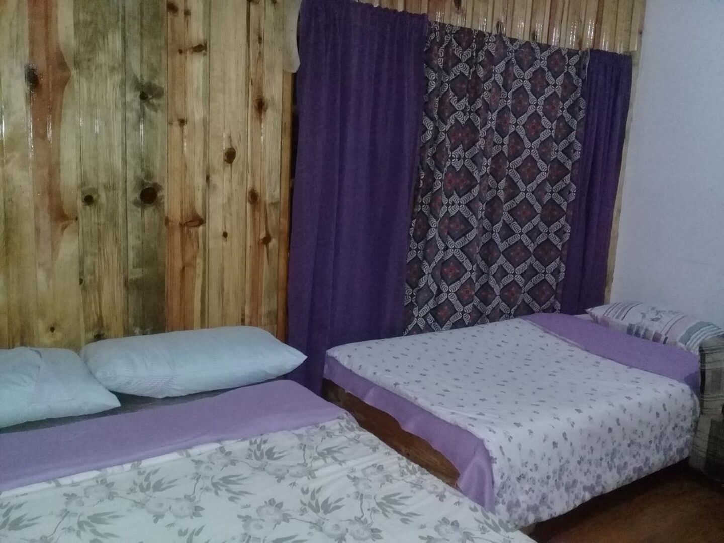 Una habitación con dos camas, paneles de madera en la pared, cortinas moradas y un cortinaje estampado.