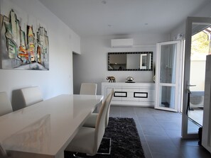 Un comedor moderno con una mesa y sillas blancas, una alfombra negra, un mueble blanco con objetos decorativos y un espejo grande encima del mueble.