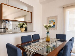 Un coin repas avec une table à plateau en verre, des chaises bleues, un vase de fleurs, un tableau accroché au mur, et un espace cuisine comprenant un évier et des placards.