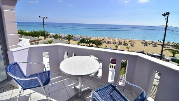 Un balcon avec une table et des chaises, donnant sur une plage parsemée de parasols et de transats.
