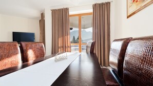 Ein Hotelzimmer mit einem langen Esstisch, zwei gemusterten Sofas, einem Fernseher und einem großen Fenster mit Vorhängen.