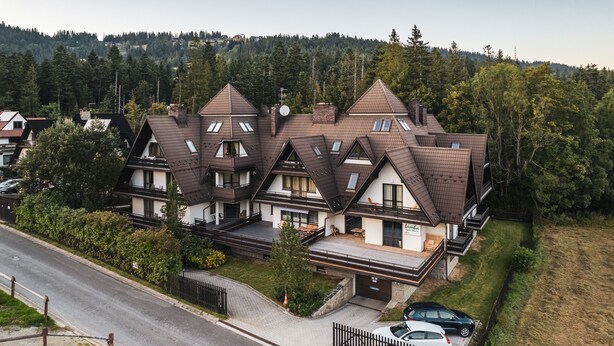 Ein mehrgeschossiges Haus mit geneigtem Dach, mehreren Balkonen und einer Auffahrt mit parkenden Autos.