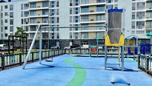 A playground with swings and climbing structures, surrounded by a fence, with a multi-story building in the background. - Azores Paim Apartment T1 (Ponta Delgada)